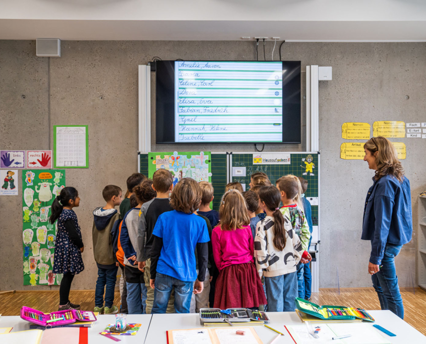 19 Schüler stehen interressiert vor der Tafel über der ein großes Videoboard hängt