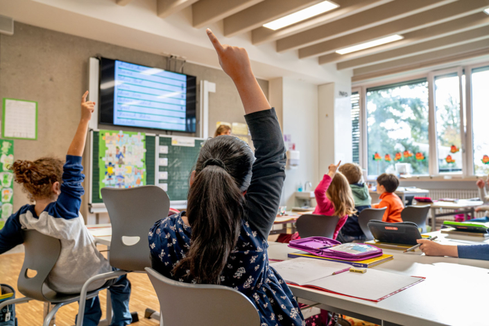 Einige Schülerinnen mit Blick auf die Tafel und die Lehrerein melden sich